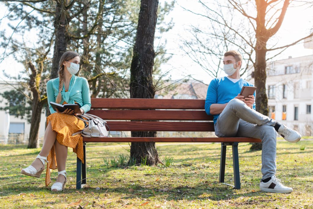 Woman and man wearing face masks and socially distancing on a park bench while reading